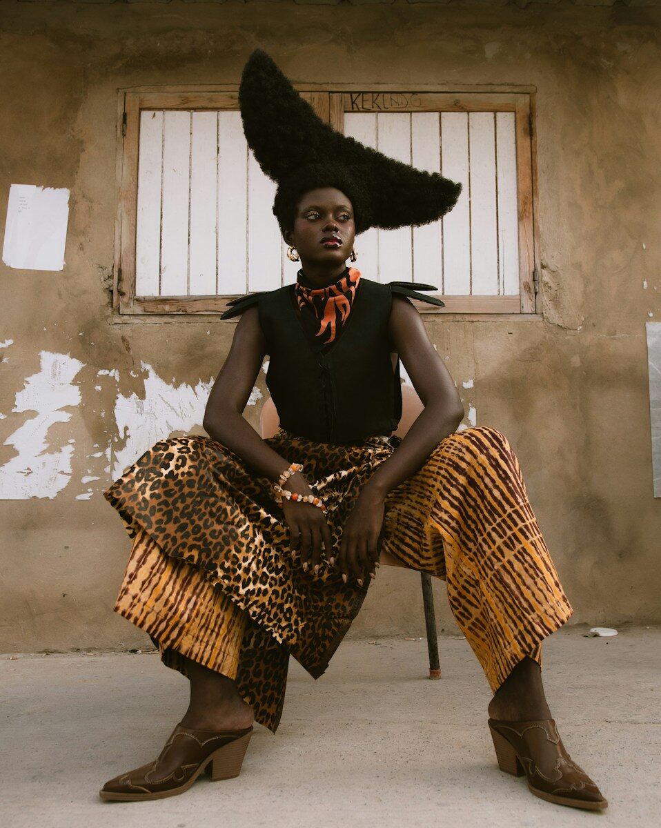 A person with a tall sculptural hairstyle wearing a black vest and patterned trousers sitting on a chair